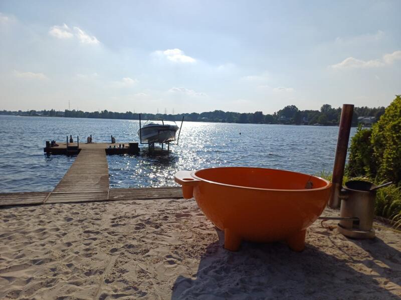 Oranje Dutchtub met houtkachel op een zandstrandje aan het meer, met een steiger en een boot